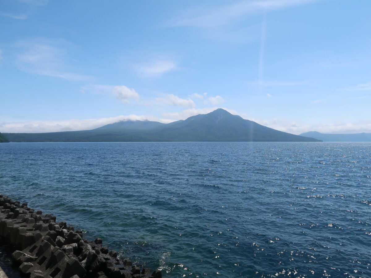 支笏湖越しの樽前山(左奥)。手前の尖った山は<ruby>風不死岳<rt>ふっぷしだけ</rt></ruby>。これらの山の間の谷のひとつが、苔の回廊だ