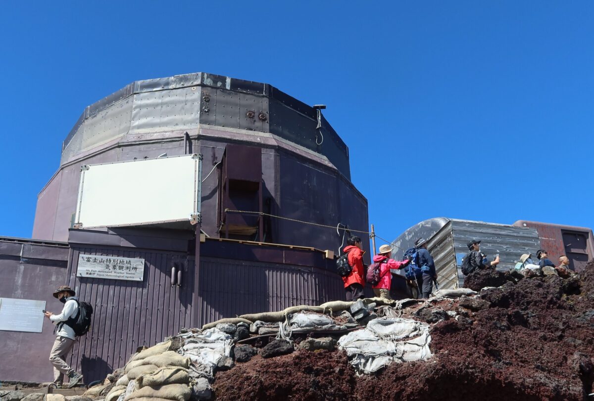 剣ヶ峰の山頂標のすぐ隣に残っている旧「富士山測候所」。「富士山特別地域気象観測所」という看板はそのままだが、内部には入れない。その右側には、山頂標の前で記念写真を撮るために登山者が並んでいる